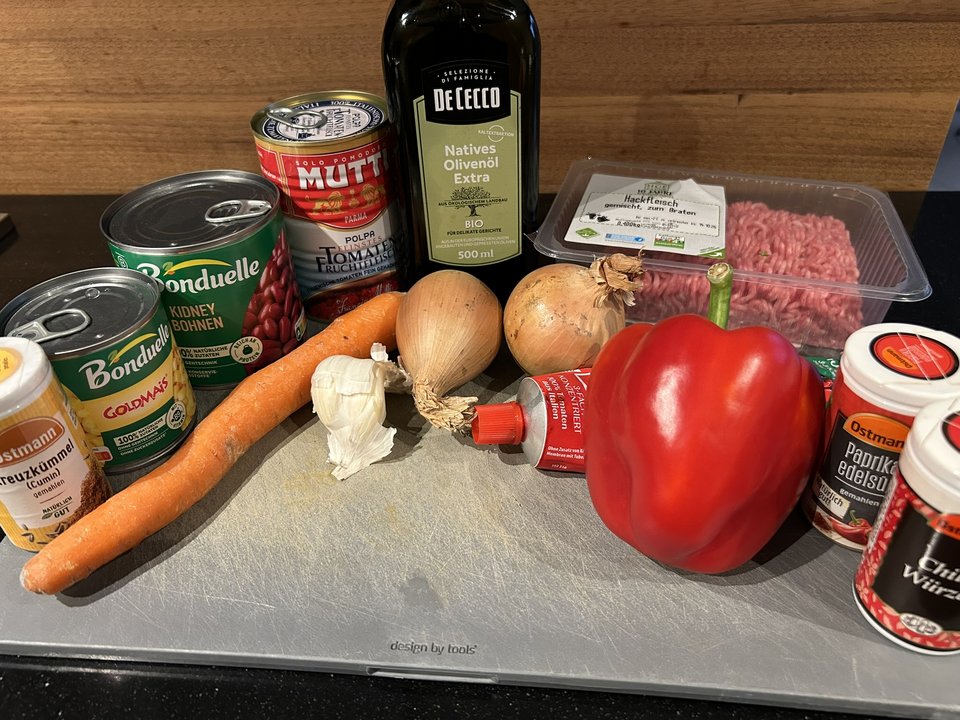 Hackfleisch, Tomaten, Kidneybohnen, Mais, Paprika, Möhre, Zwiebeln, Knoblauch, Tomatenmark, Olivenöl und Gewürze auf einem Schneidebrett