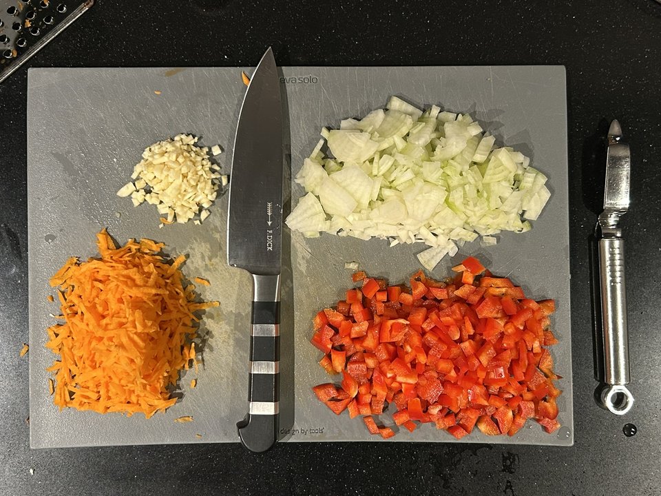 Gehackter Knoblauch, geschnittene Zwiebeln, gewürfelte rote Paprika und grob geraspelte Möhre auf einem Schneidebrett